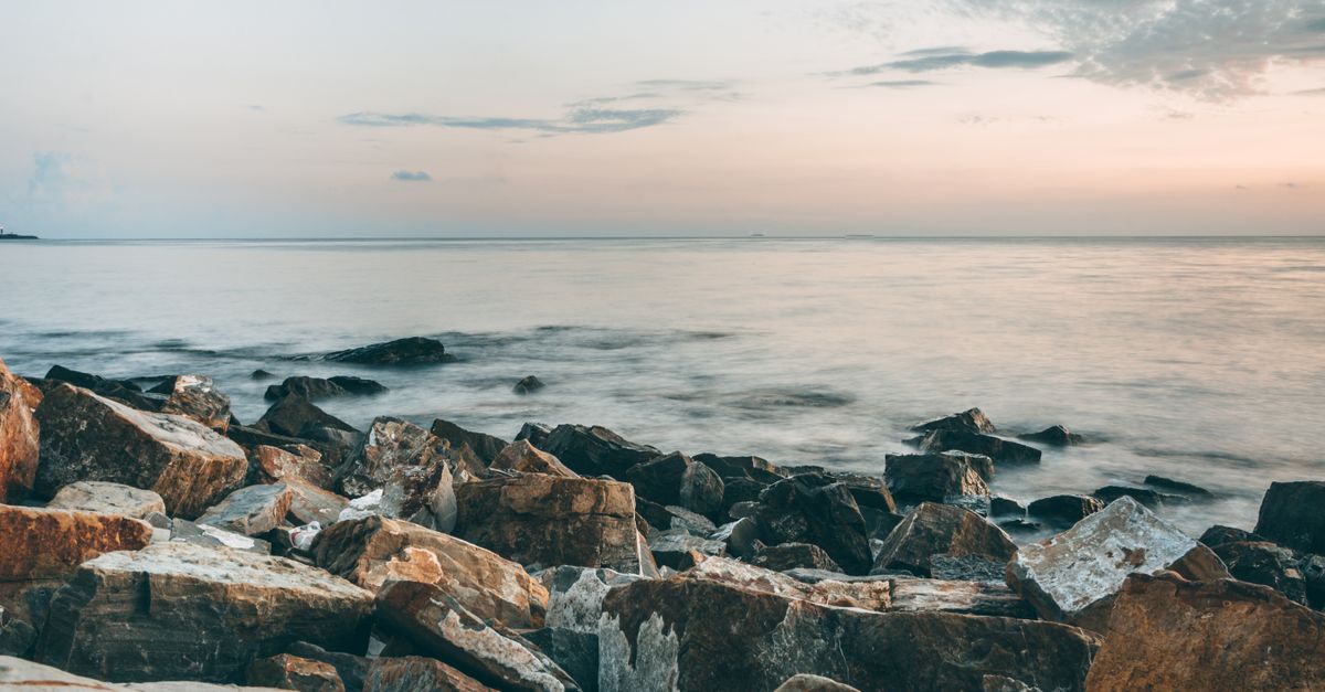 Scenic view of rocky formations on coast against rippled sea with horizon under cloudy sky at sunset