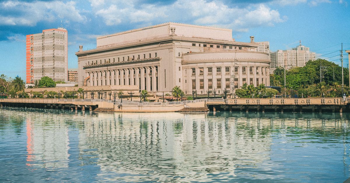 The iconic Manila Post Office building reflecting on the Pasig River under a bright blue sky.
