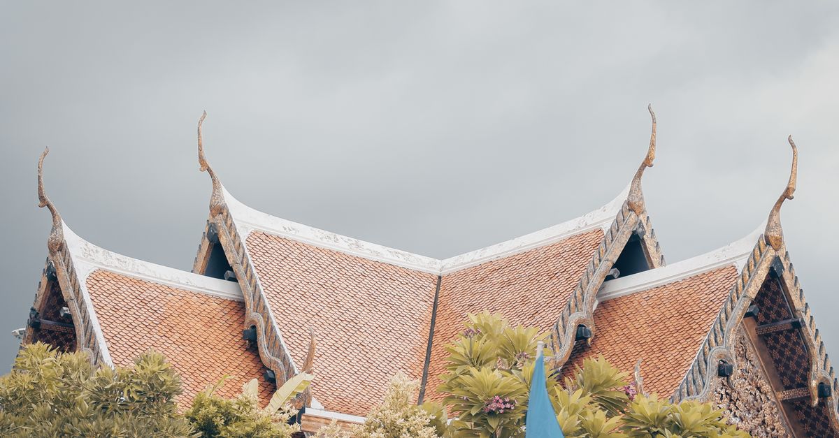 Elegant traditional Thai temple roof with a bird flying above, surrounded by lush green trees in Bangkok, Thailand.