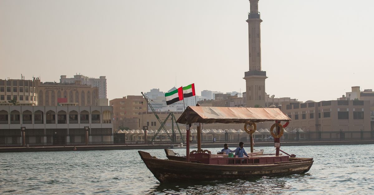 A traditional abra boat sails on Dubai Creek with a city skyline and minaret in the background.