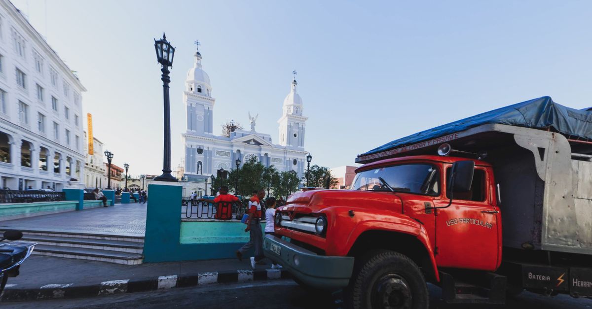 A vintage red truck parked in a bustling city square with a historic cathedral in the background.