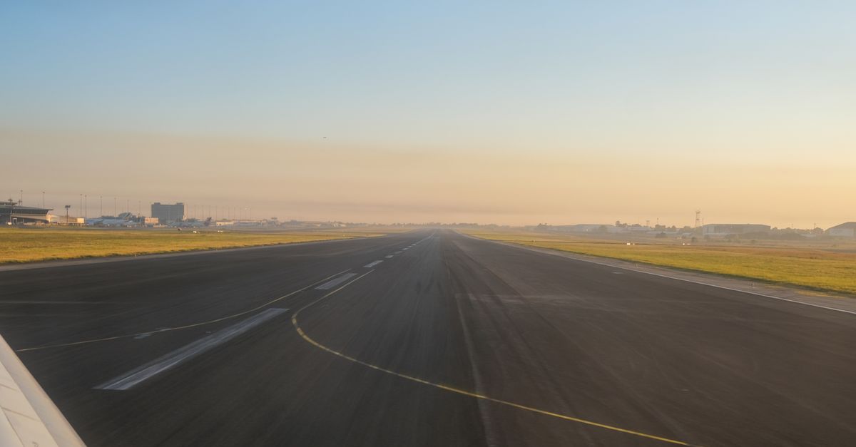 An empty airport runway at sunrise, showcasing a clear sky and distant airport buildings.