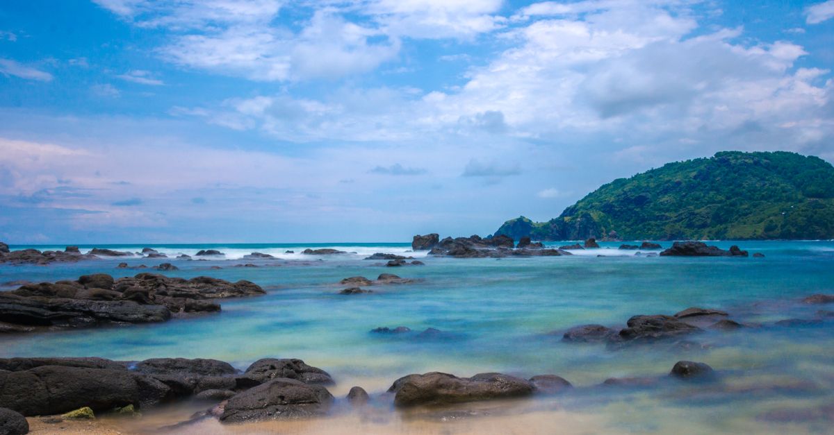 Idyllic tropical beach scene with rocky shoreline, clear blue waters, and lush hill in the background.