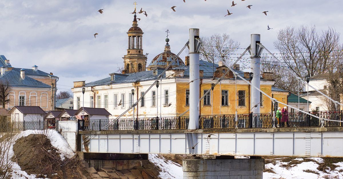 A scenic view of a historic bridge and church in winter, Торжок, Russia, with birds flying overhead.