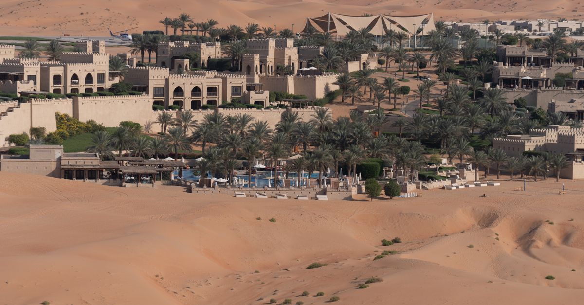 Aerial view of a luxurious desert oasis resort surrounded by vast sandy dunes and palm trees.