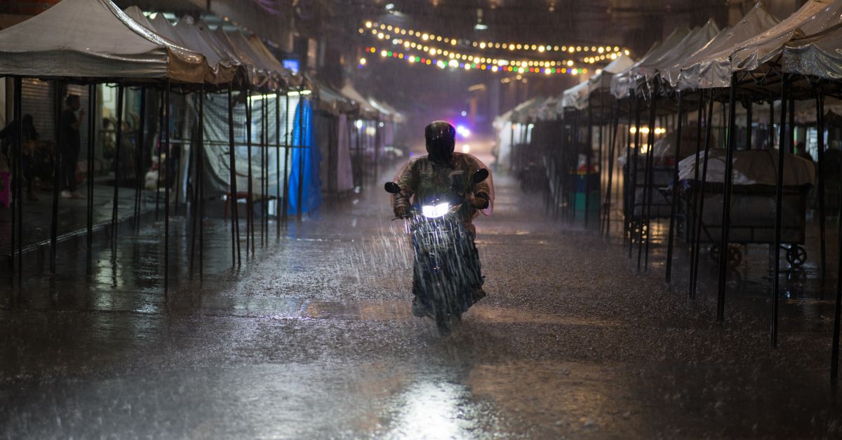 Motorcycle rider braves heavy rain in Bangkok street lined with tents and string lights at night.