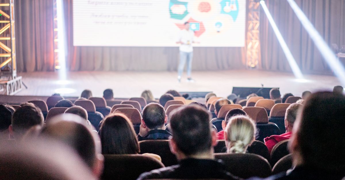 Audience attentively watching a presentation in a conference setting, with stage lights and speaker visible.