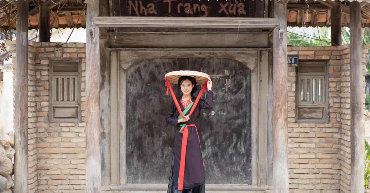 Woman in traditional Vietnamese outfit with wooden house background.