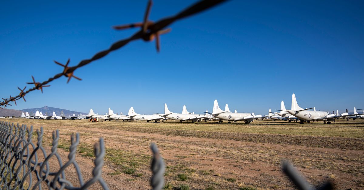 Airplane boneyard viewed through barbed wire fence under clear blue sky.