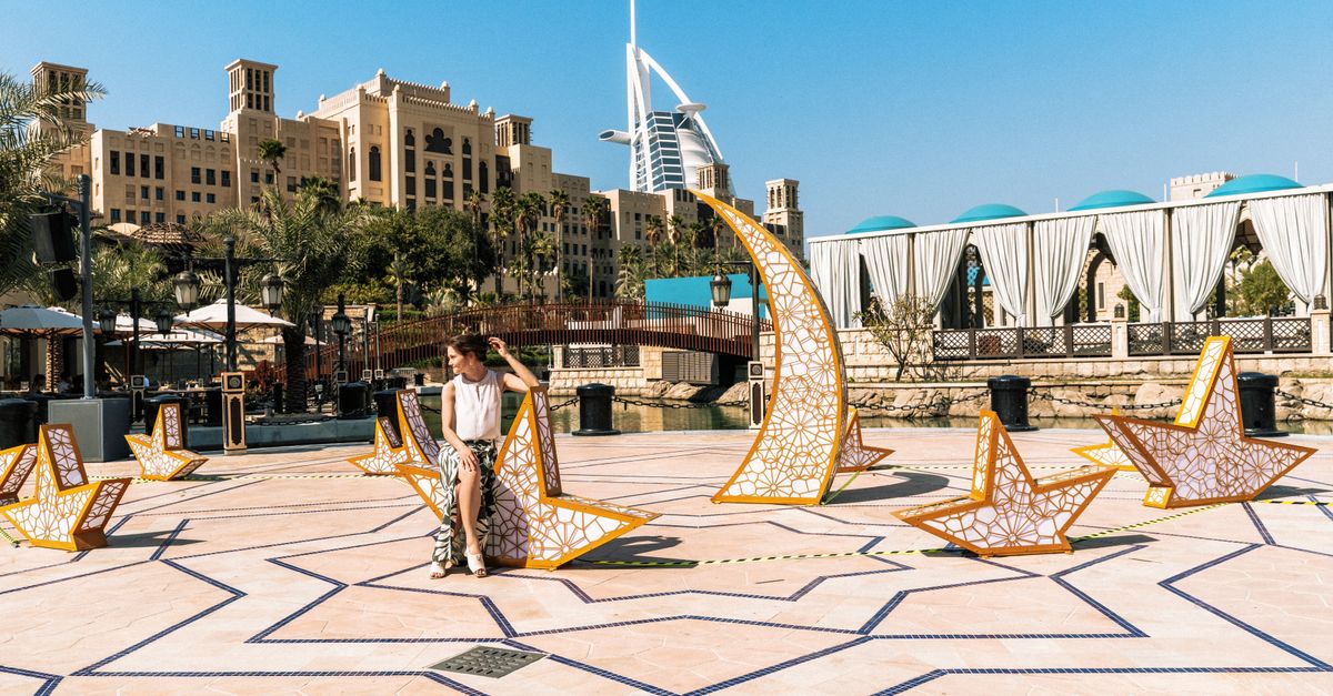 A woman relaxes by an artistic installation with the iconic Burj Al Arab in the background, set in Dubai's luxurious outdoor setting.