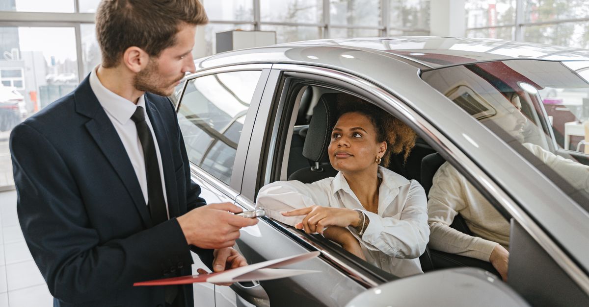 A car dealer hands keys to a woman sitting inside a vehicle in a car showroom.