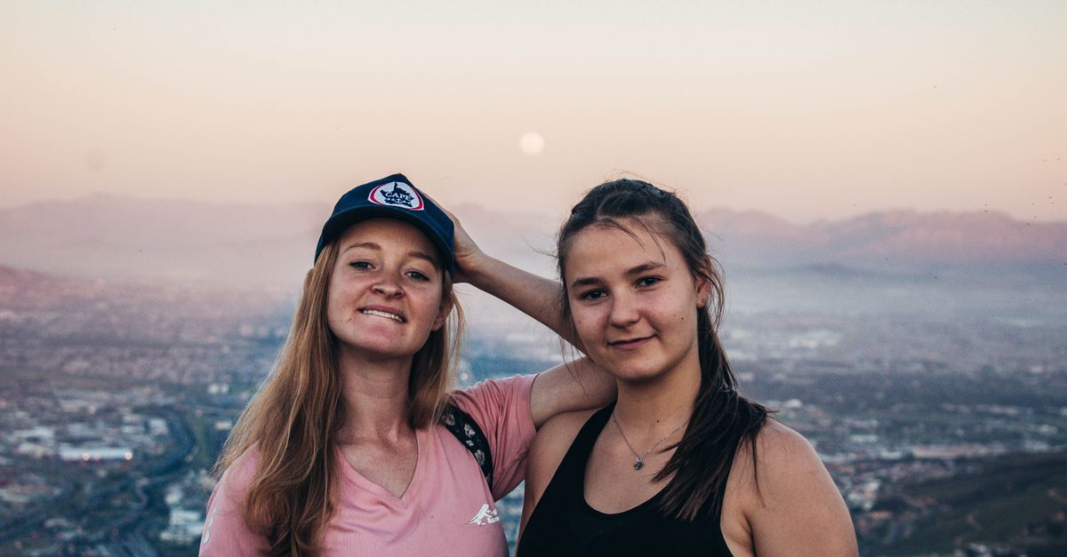 Two young women pose together with a scenic mountain view at sunset, showcasing friendship and travel.