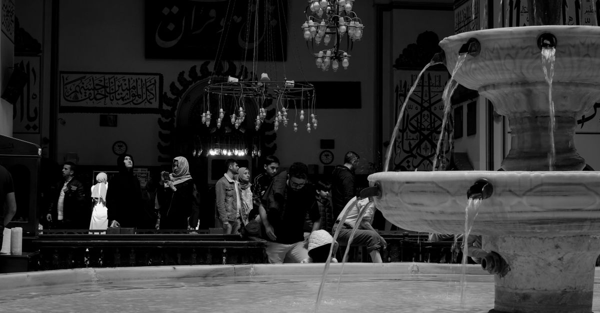 Black and white image of a mosque interior with fountain and people in Bursa, Türkiye.