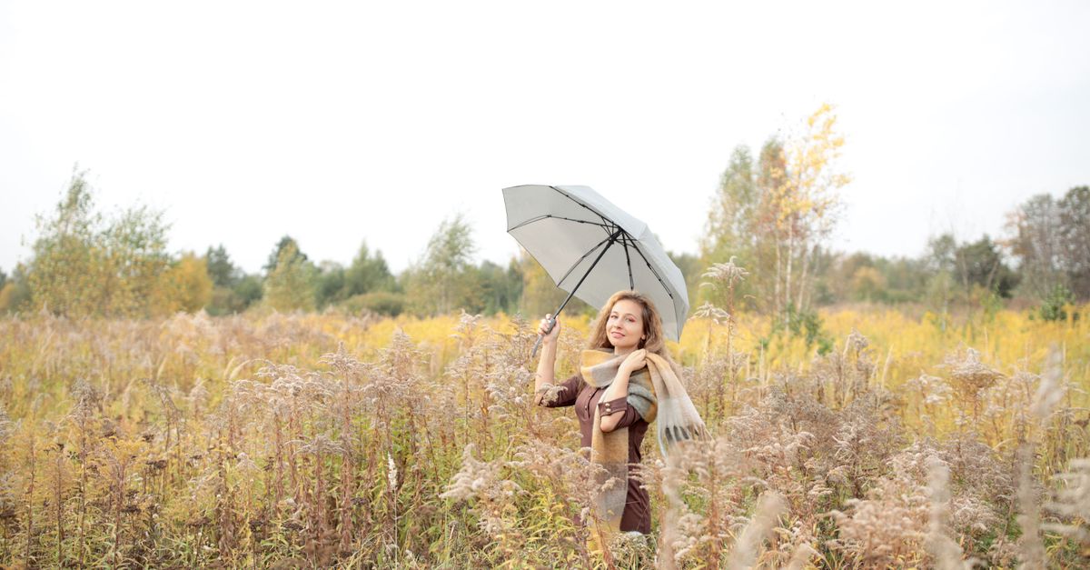 A woman stands in a fall field holding an umbrella, surrounded by autumn foliage.