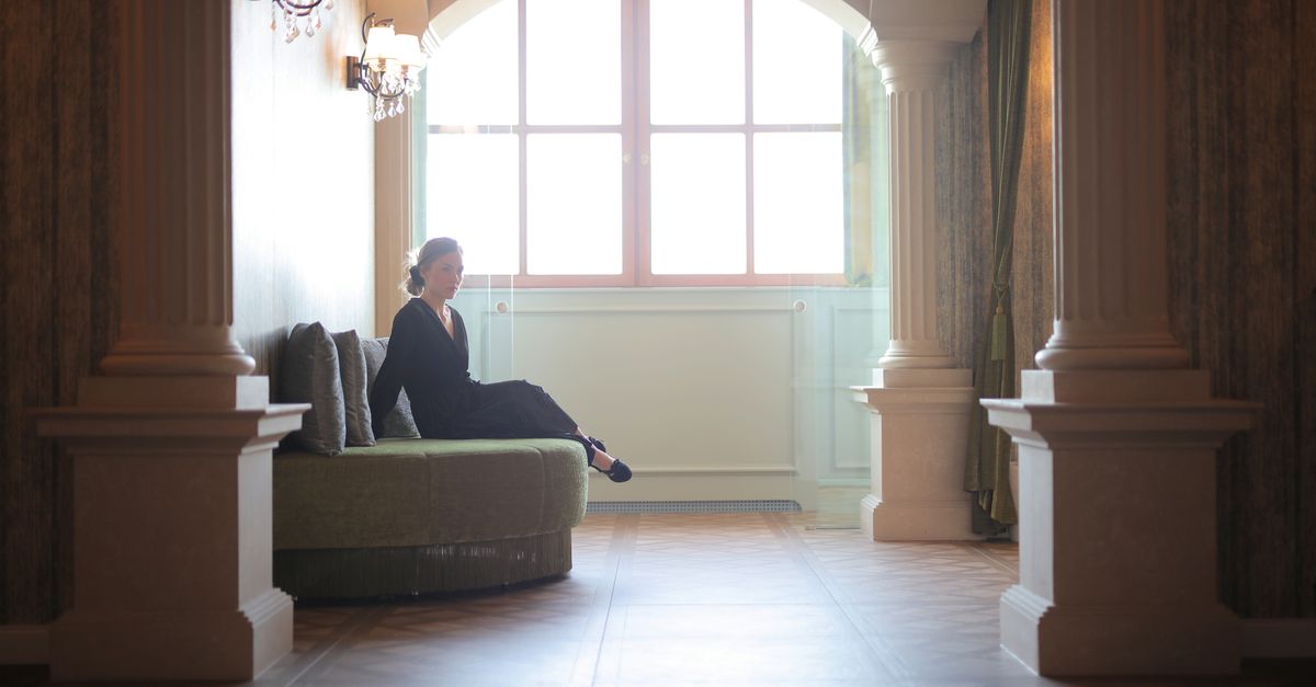 A woman in a black dress sits elegantly in a classic lobby framed by architectural columns.