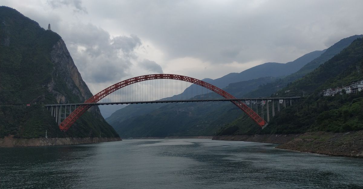 Stunning red arch bridge over a wide river with lush mountains beneath a cloudy sky.