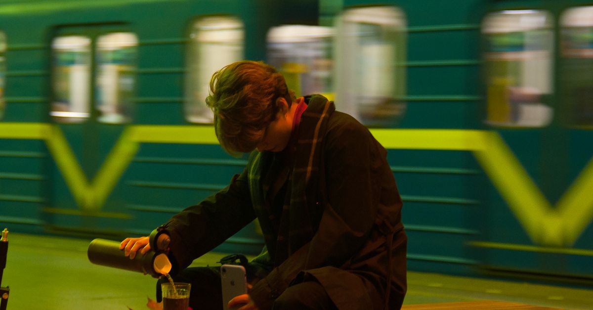 Woman pours drink from tumbler at train station, St. Petersburg. Vibrant metro in background.