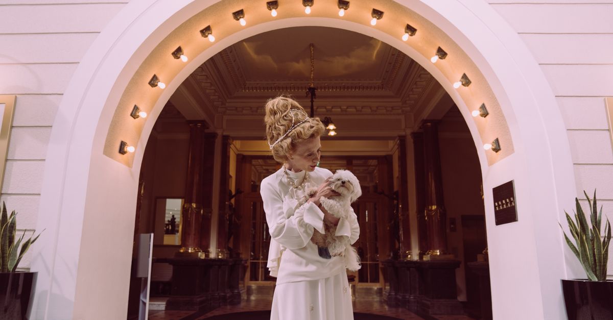 A senior woman elegantly dressed, holding a small dog in a luxurious hotel lobby.
