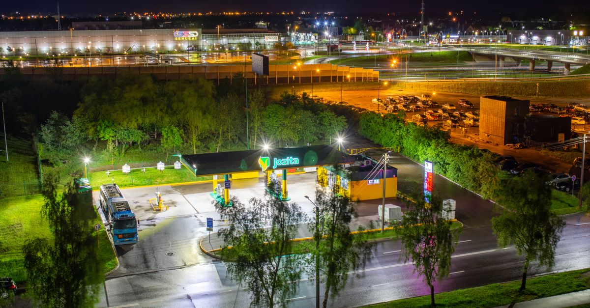 A brightly lit gas station at night, surrounded by a vibrant cityscape and highway.