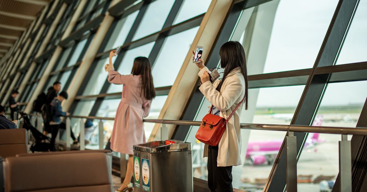 Two young women taking selfies at a modern Shanghai airport terminal with large windows.